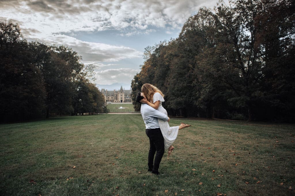 A couple embraces on lawn in front of the Biltmore House. The houses is in the background, surrounded by trees under blue skies with fluffy white clouds.