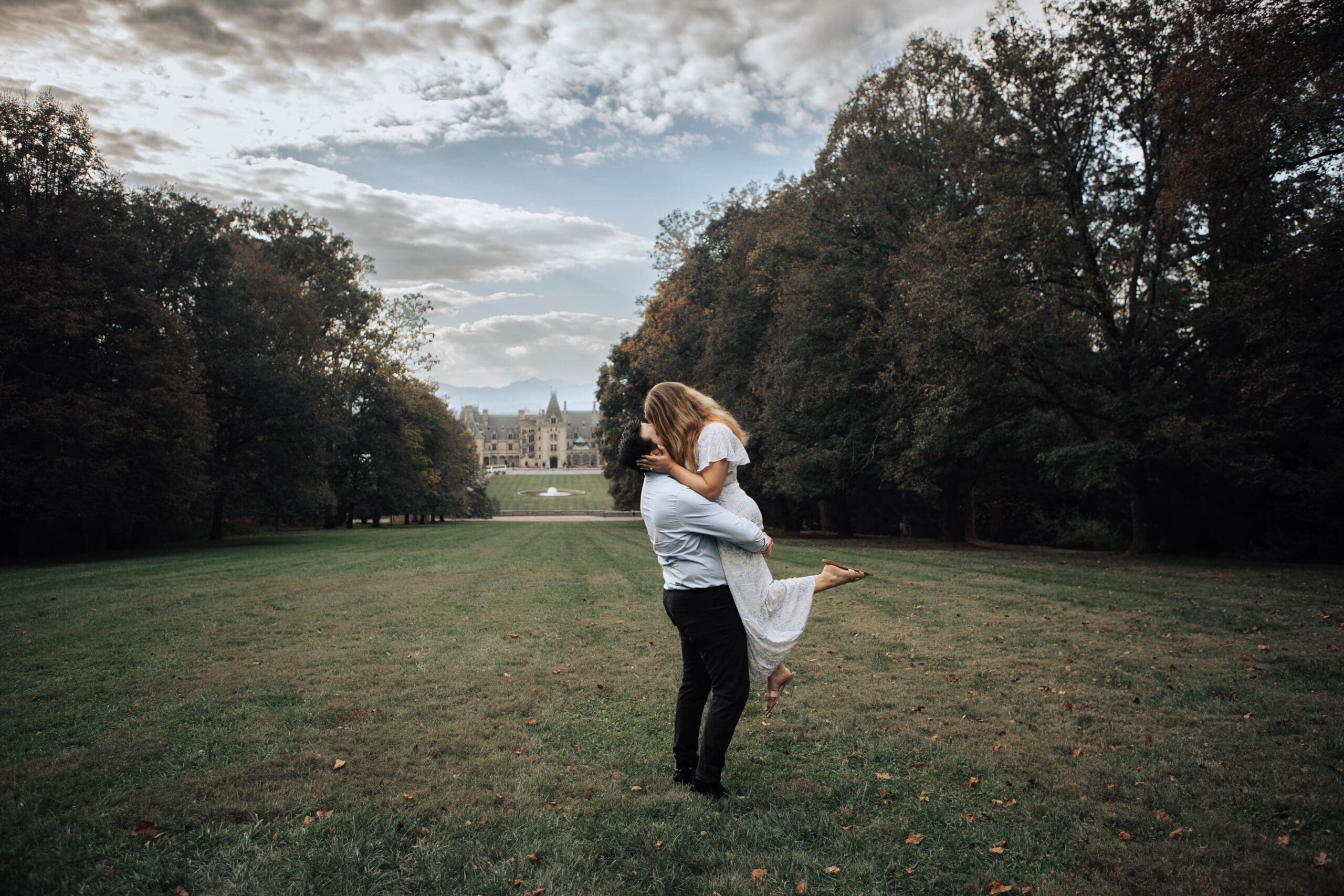 Couple embraces on lawn of the Biltmore estate. The Biltmore House is in the background under blue skies with fluffy white clouds.