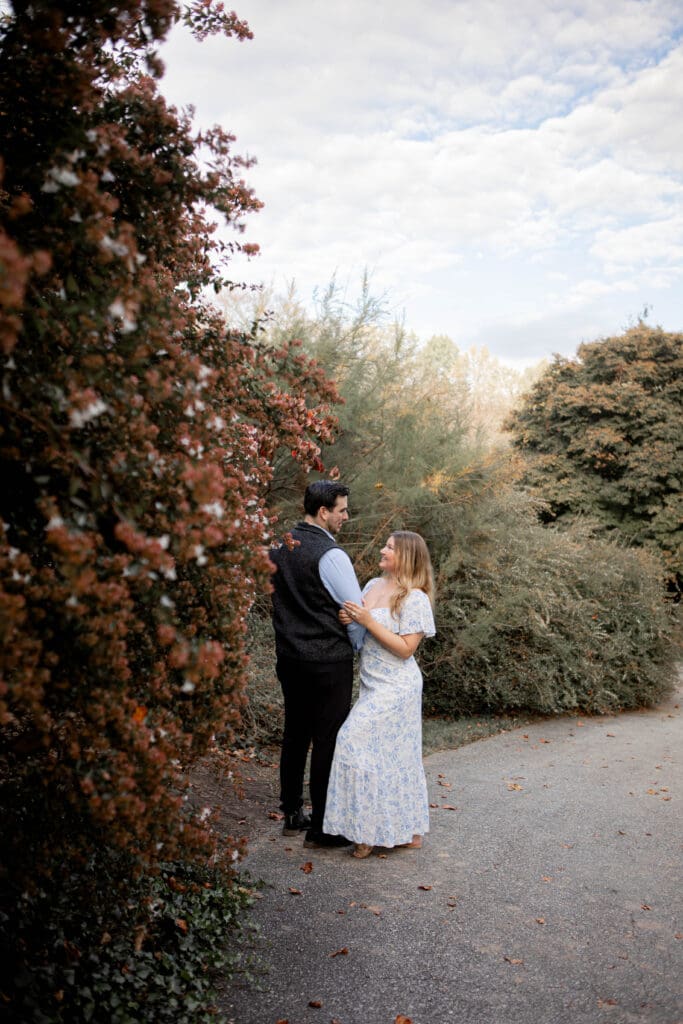 A man and woman embrace and look at each other standing near trees that are turning yellow and orange. She is wearing a blue and white dress.
