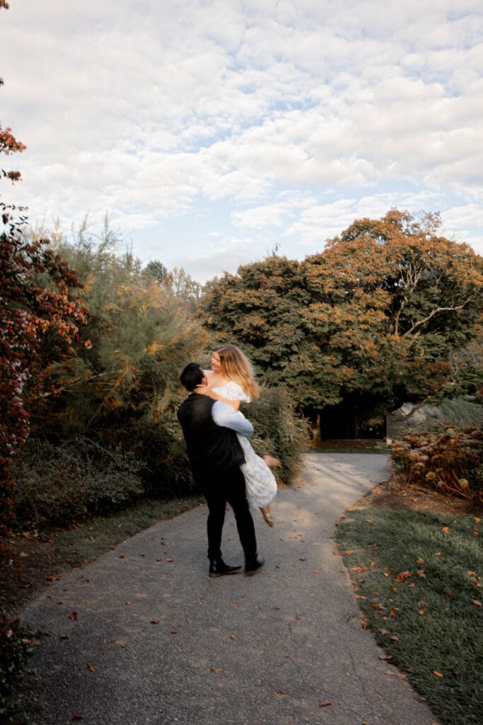 A man is swinging a woman around in a passionate embrace. They are surrounded by trees with leaves slowly turning red and orange.