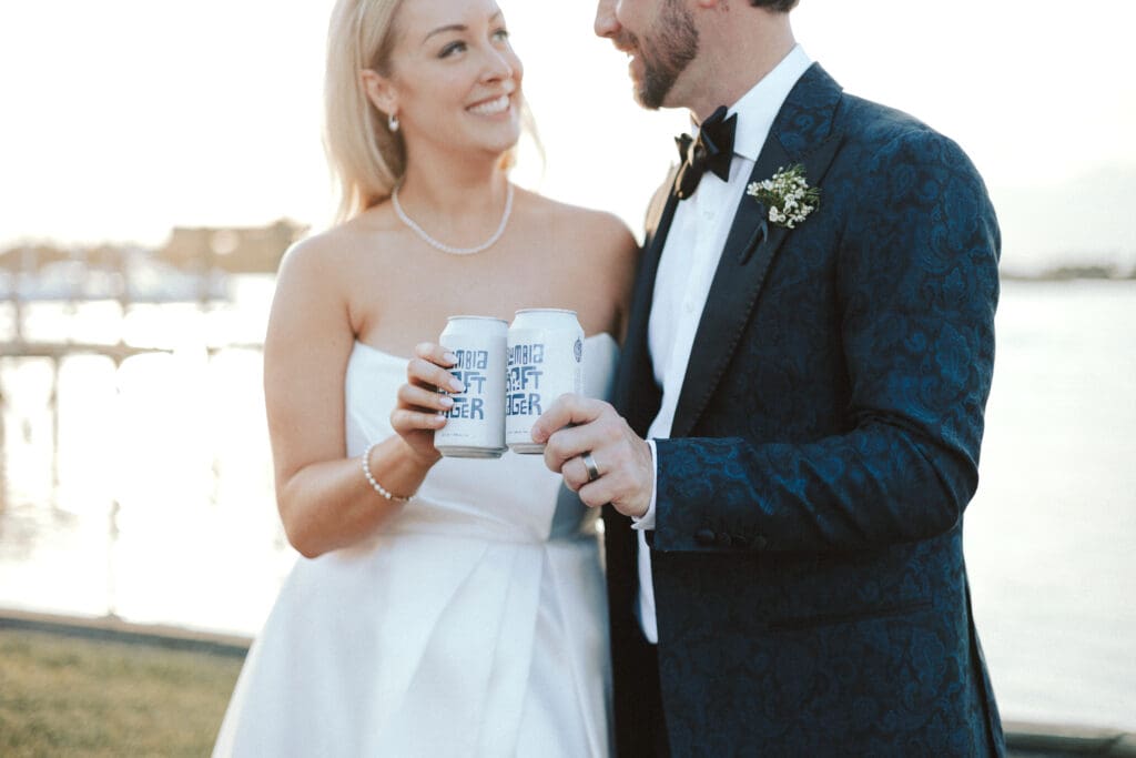 A bride and groom cheers with craft beer cans standing in front of a body of water at sunset.