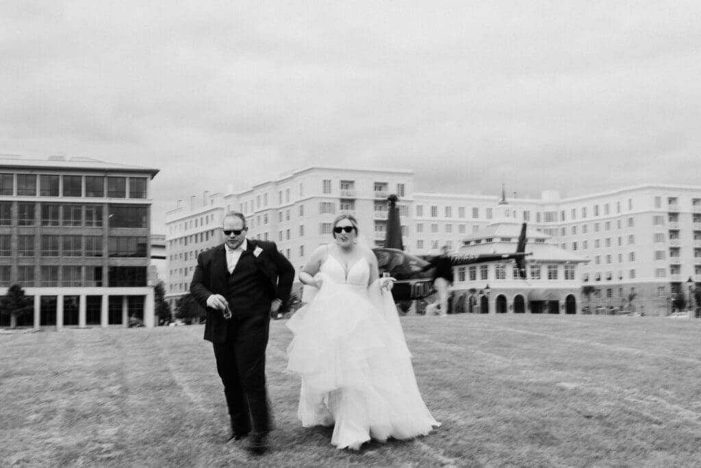 A bride and groom walk toward the camera, wearing sunglasses, holding beers, with a helicopter behind them.