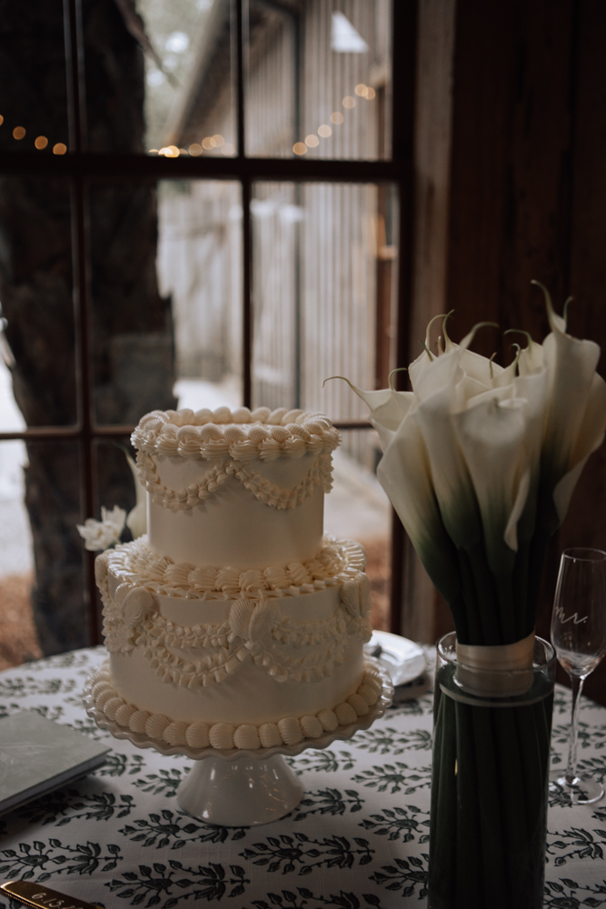 A beautiful white wedding cake next to calla lilies on a green and white table cloth. The window in the background has string lights.