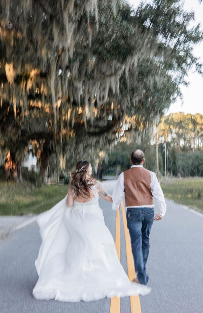 A bride and groom run down the road. Her hair flows in the breeze. They're surrounded by wispy trees.