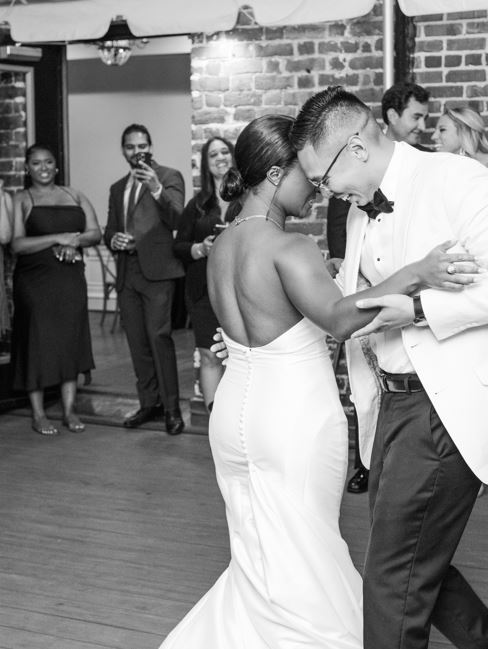A bride and groom dance. The photo is black and white. People watch in the background.