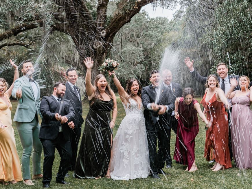 A bride and groom and several of their colorfully dressed friends spray champagne and laugh.