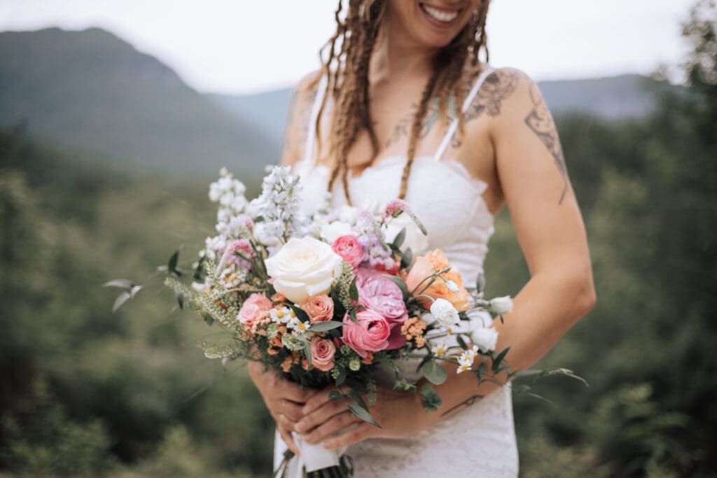 A bride with tattoos holding a bouquet and smiling with mountains behind her.