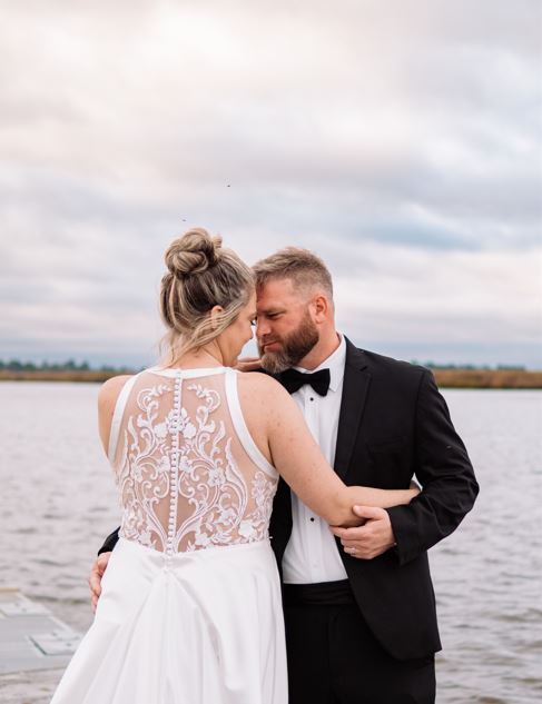 A bride and groom embrace on a dock. They're surrounded by water and fluffy clouds.