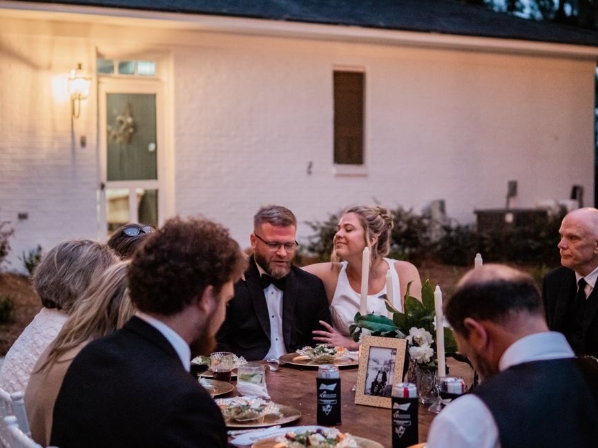 A bride and groom sit at a table with a handful of other people looking at each other. 