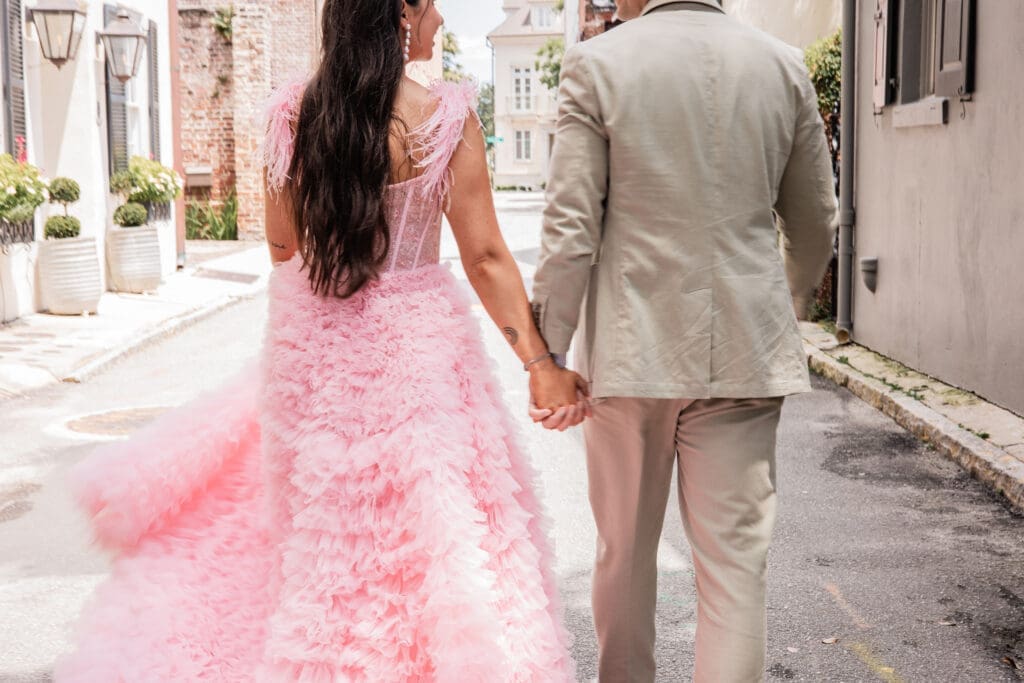 A bride wearing a fluffy pink gown and a groom in a tan suit walk down the street holding hands and smiling.
