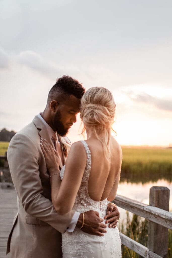 A bride and groom embrace. They're standing on a dock with the marsh behind them. The sun is setting.