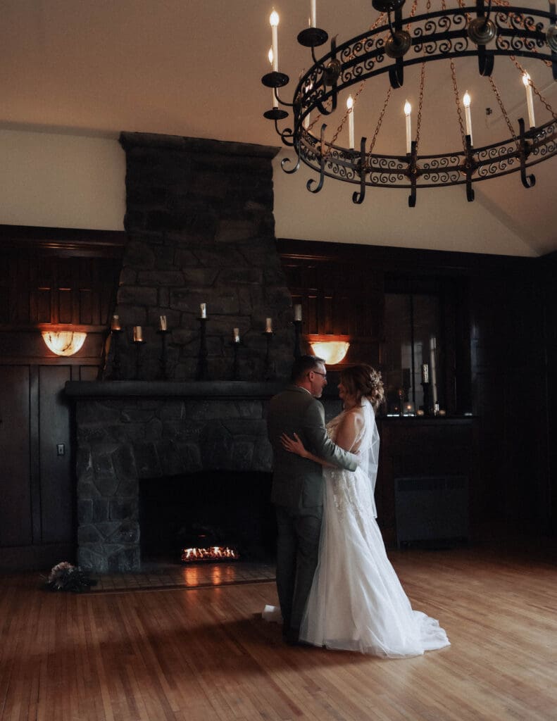 A bride and groom dance in front of a grand fireplace surrounded by candles under a large chandelier.