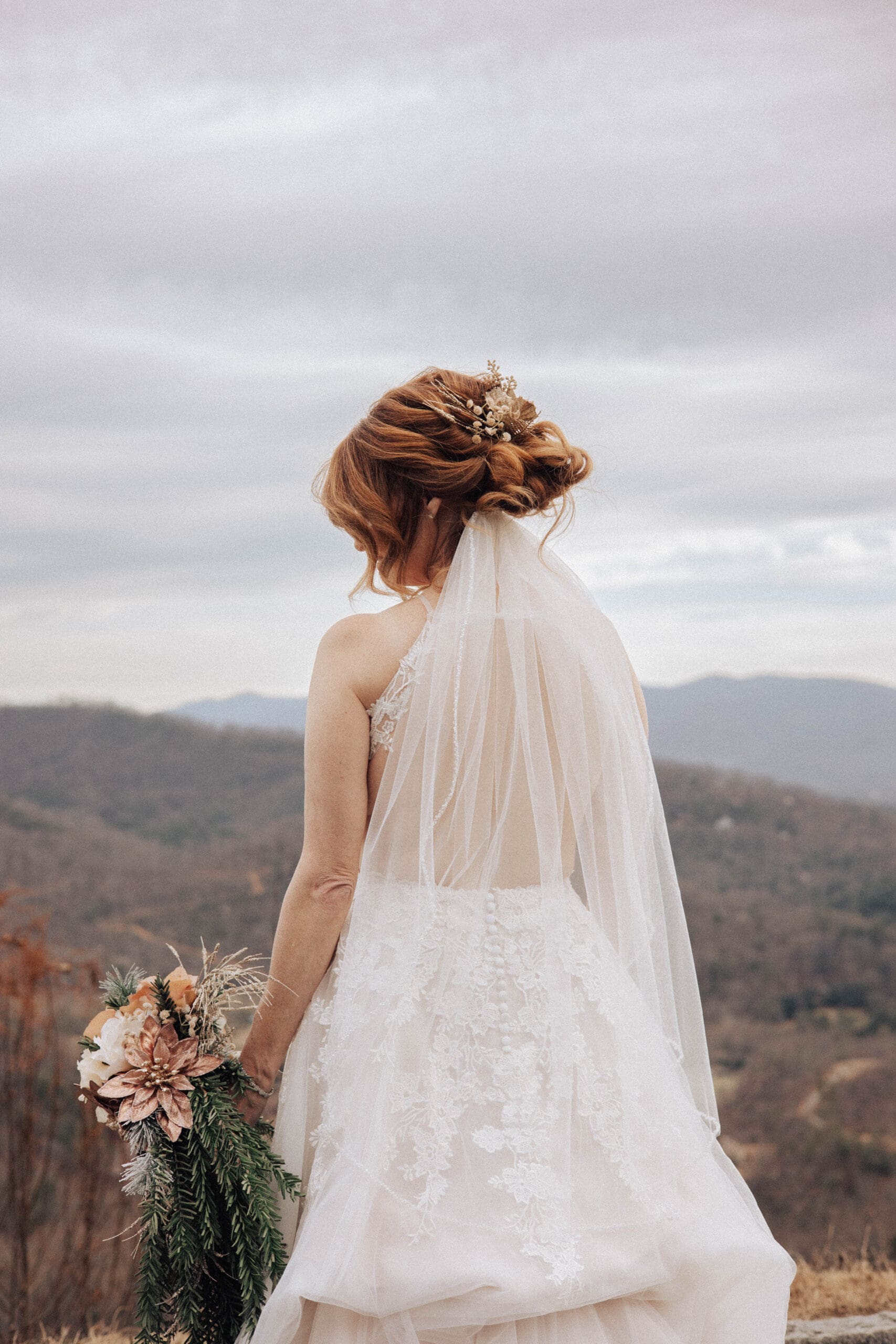 A bride stands looking over the Asheville mountains, holding a bouquet at her side, a long veil over her shoulder.