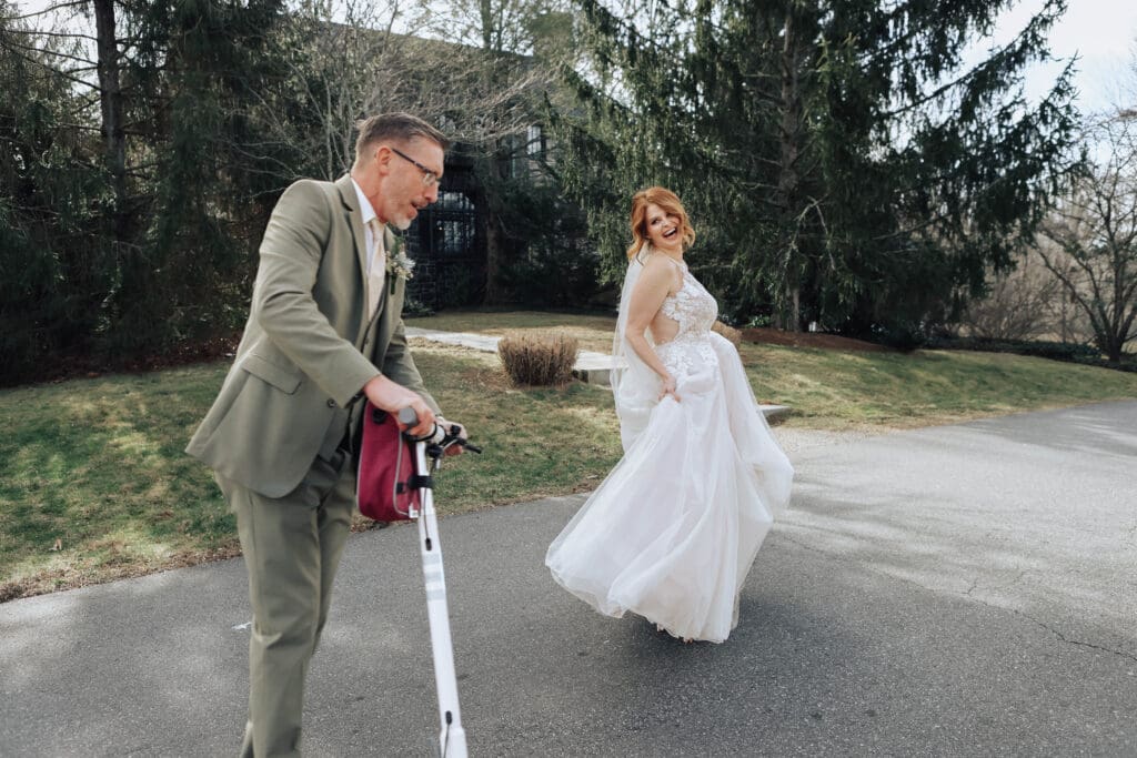 A bride in a white gown laughs as her groom zips around her on a scooter.