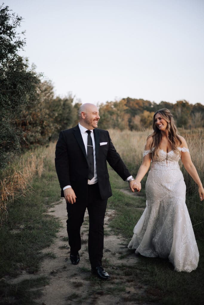 a bride and groom walk down a path in the woods, holding hands and laughing while looking at each other.