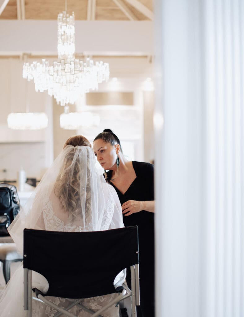 A bride sits in a chair while a makeup arts wearing all black does her makeup. There is a crystal chandelier behind them. 