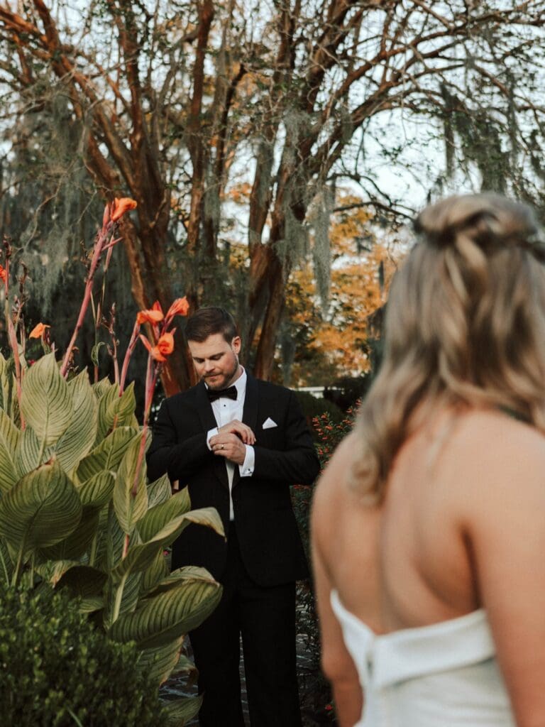 A bride watches as her groom adjusts his suit. They stand a grove with red flowers.