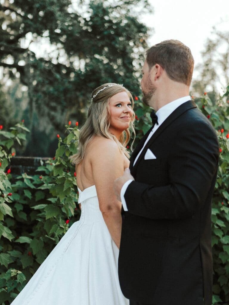 A bride and groom smile at each other surrounded by red flowers.